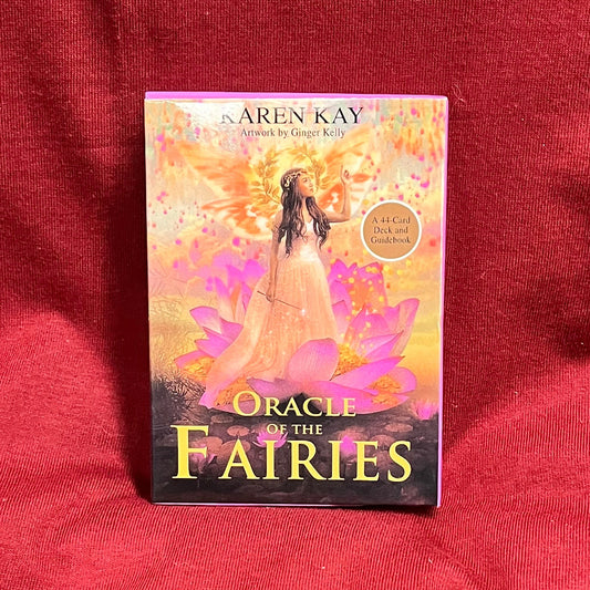 A boxed card deck titled "Oracle of the Fairies" by Karen Kay stands upright on a red fabric surface. The box features fantasy artwork with pink, orange, and purple tones, with a beautiful fairy. 
