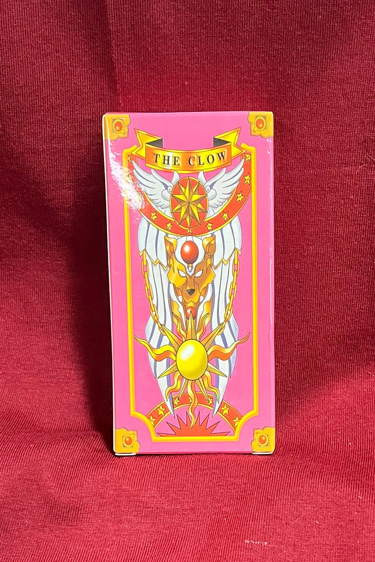 A pink tarot card deck with a winged figure and gold details stands upright on a draped red fabric surface.