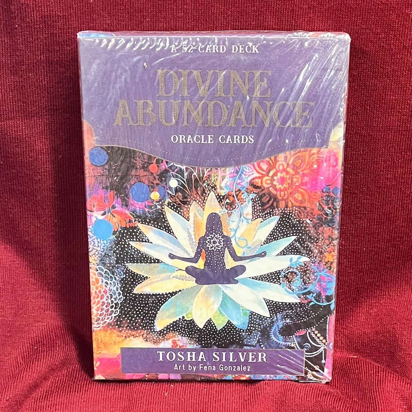 A boxed deck of oracle cards titled 'Divine Abundance Oracle Cards' by Tovia Silver, wrapped in clear plastic and standing upright on a red fabric surface.