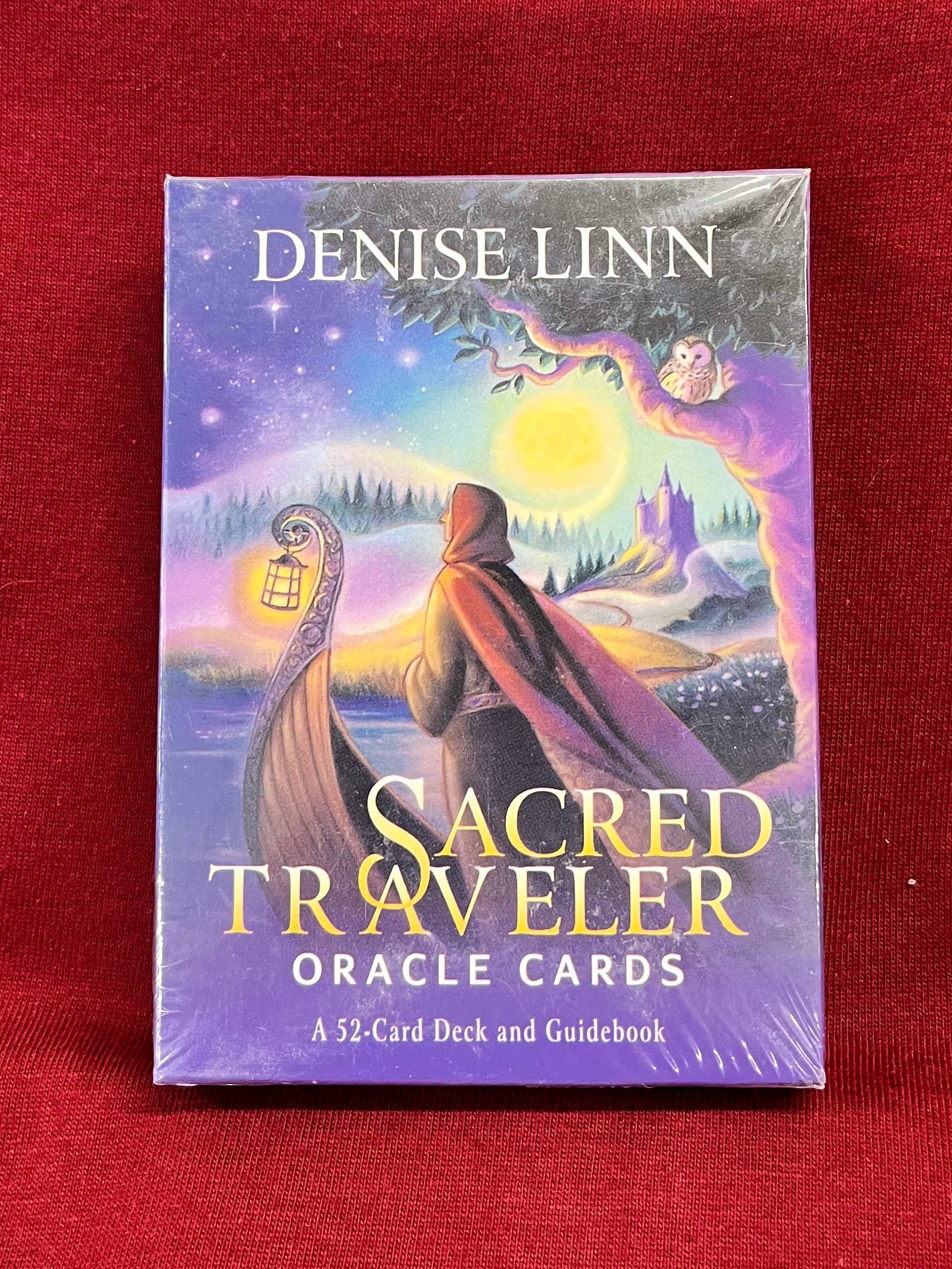 A boxed card deck titled "Sacred Traveler Oracle Cards" by Denise Linn, standing upright on a red fabric surface. On the deck, a woman stands on our in front of a boat, with a lake and a castle in the background.  A night sky full of stars and a full moon casts light on the scene.