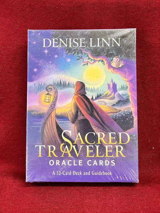 A boxed card deck titled "Sacred Traveler Oracle Cards" by Denise Linn, standing upright on a red fabric surface. On the deck, a woman stands on our in front of a boat, with a lake and a castle in the background.  A night sky full of stars and a full moon casts light on the scene.