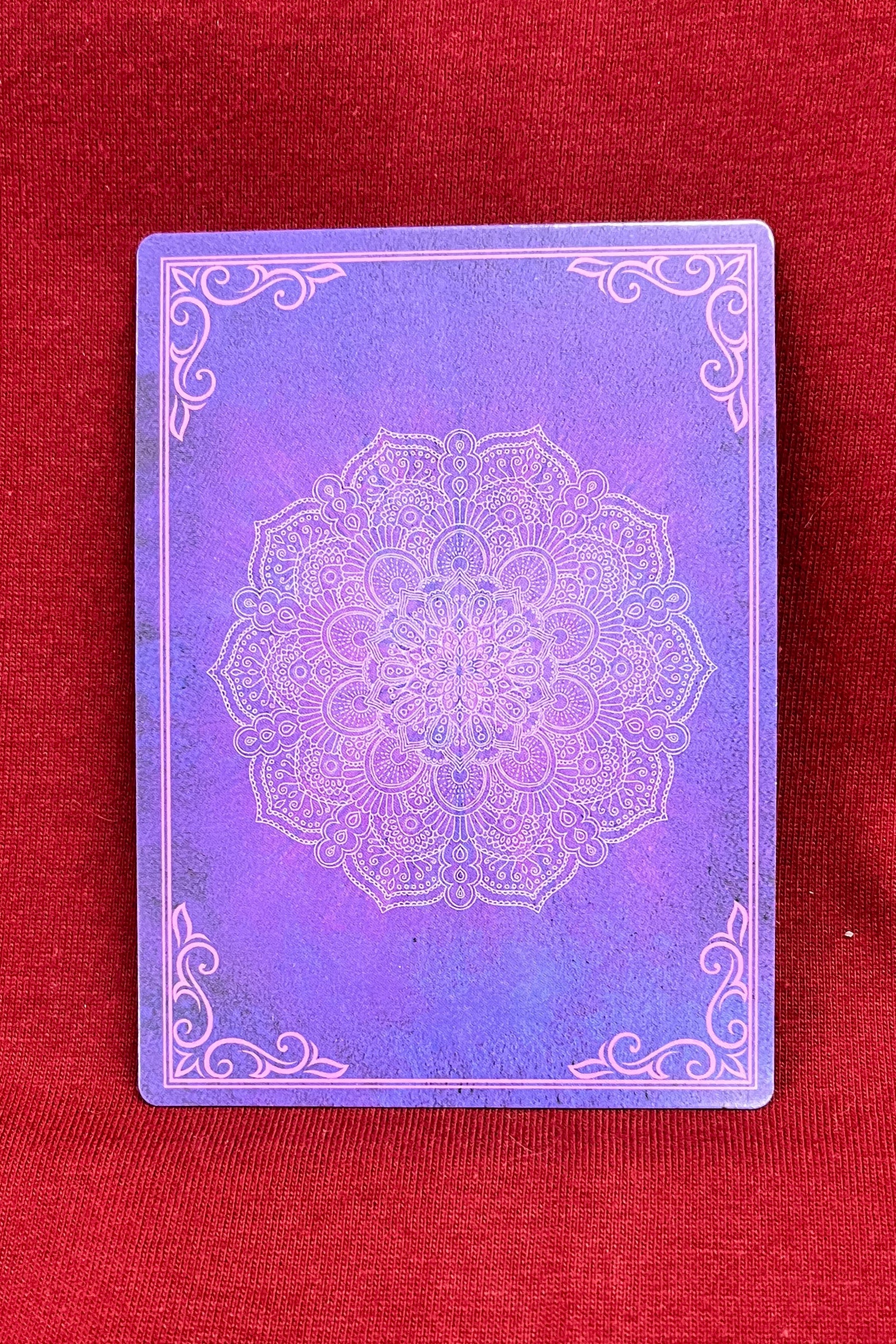 A square purple card with a white mandala design in the center and decorative corner flourishes, resting on a textured red fabric background.