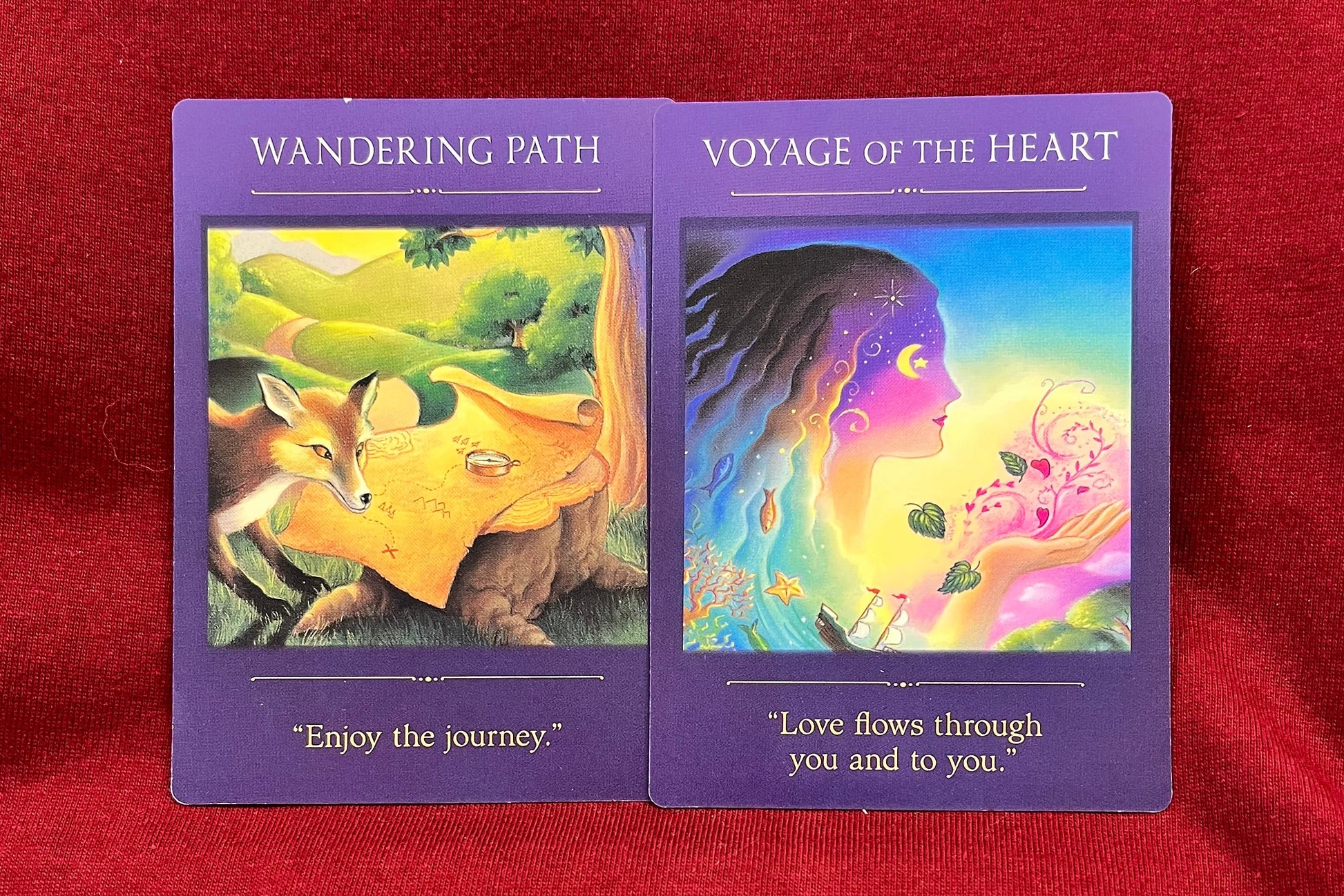 Two tarot oracle cards titled 'Wandering Path' and 'Voyage of the Heart' displayed side-by-side on a textured red fabric background. The cards feature purple borders and colorful fantasy illustrations.