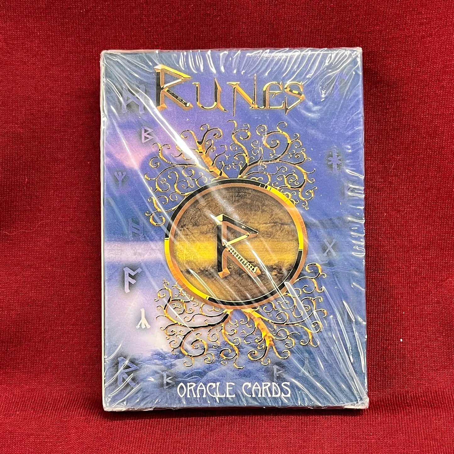 A boxed deck of 'Runes' Oracle Cards, still sealed in plastic wrap, displayed on a red fabric surface.
