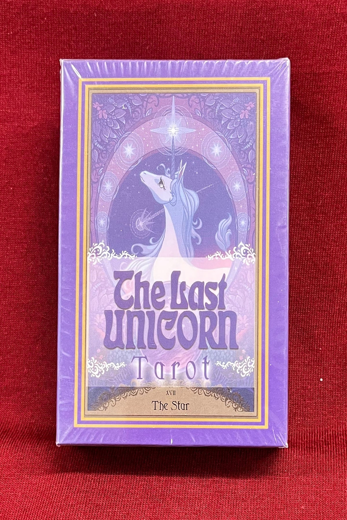The Last Unicorn Tarot deck box standing upright on a red fabric surface, with a purple unicorn illustration and the title 'The Last Unicorn Tarot' visible on the front.