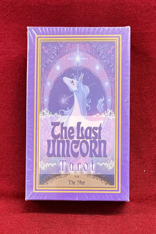 The Last Unicorn Tarot deck box standing upright on a red fabric surface, with a purple unicorn illustration and the title 'The Last Unicorn Tarot' visible on the front.