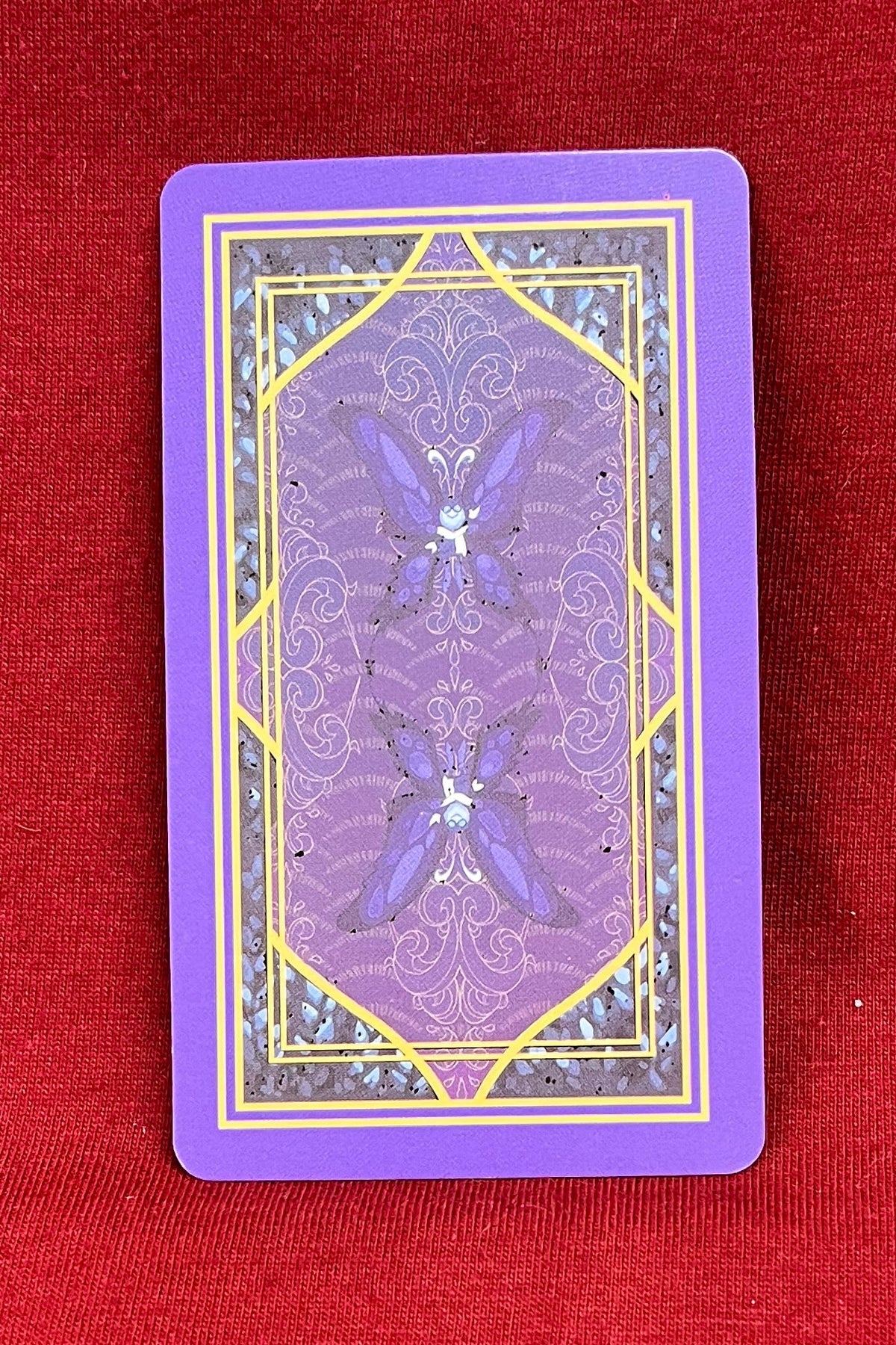 A purple oracle card back with gold and blue decorative patterns lies on a textured red fabric surface.