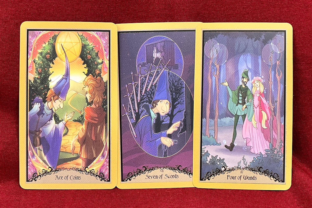 Three illustrated tarot cards displayed upright on a red fabric surface. The left card shows a figure in a blue cloak beside a woman in a red dress in front of a glowing light, the middle card depicts a dark figure with a staff under an oval, and the right card shows a man dressed in green walking beside a woman in Pink.