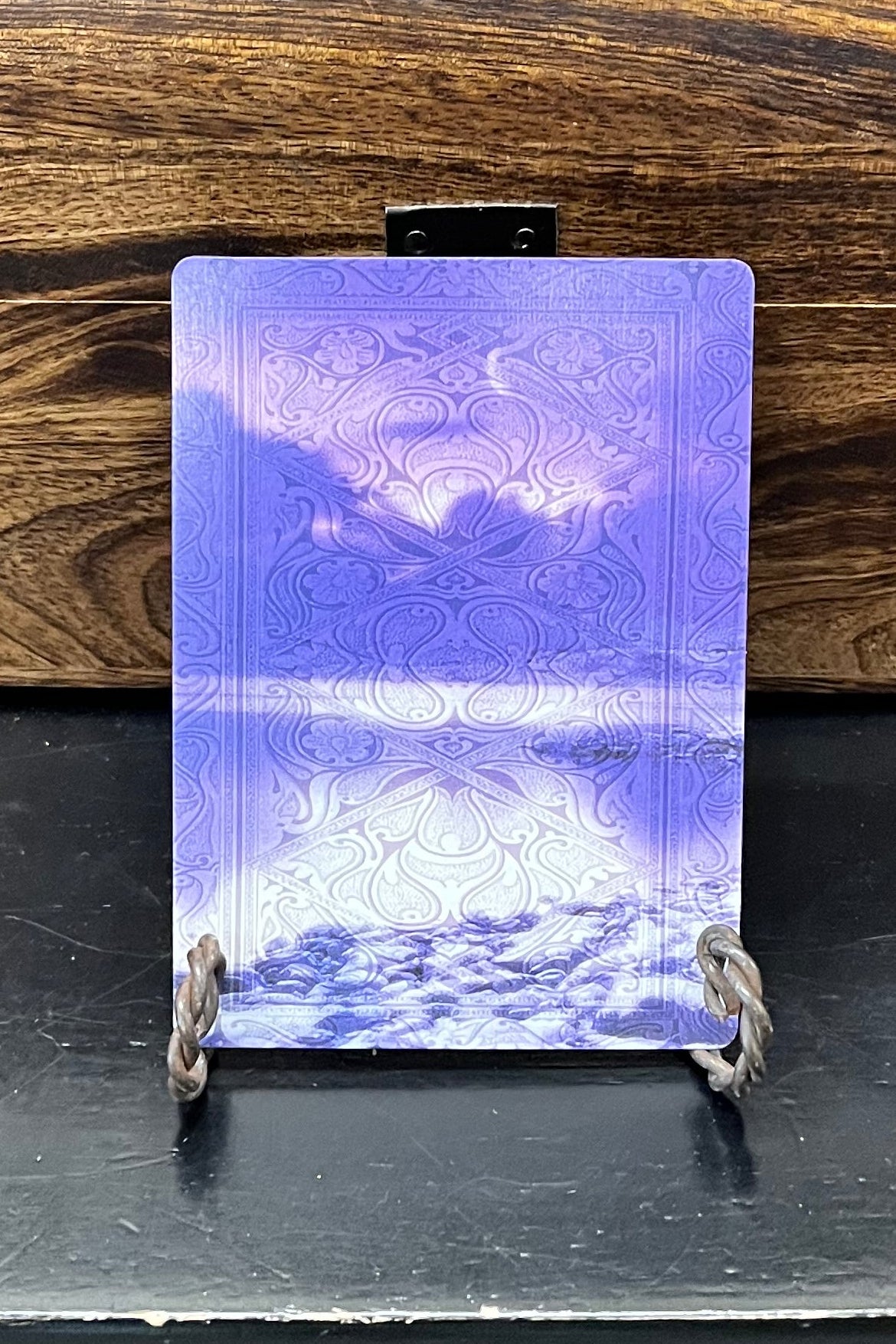 A small rectangular card or print with a purple and blue landscape scene, displayed upright on a small stand in front of a wooden chest on a black table.