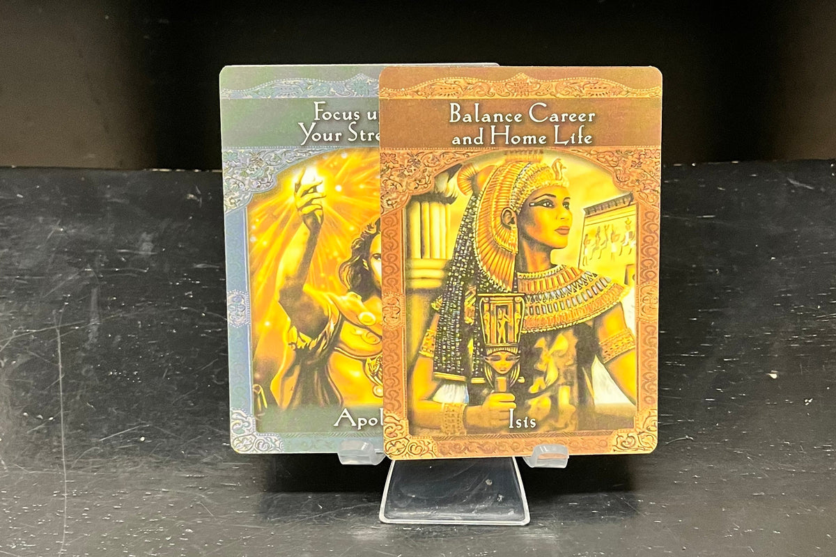 Two illustrated oracle cards standing upright on a clear display stand on a dark tabletop. The left card has a green-gray background with a yellow figure, and the right card has a warm brown background with an ancient Egyptian-style figure.