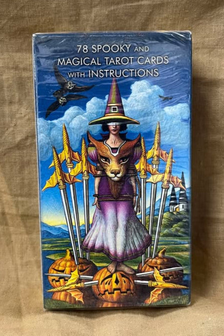 Tarot card set back shows a witch wearing the face of a cat as a breastplate, surrounded by swords and walking on a jack-on-lantern with a sad face pierced by two swords.  Two birds fly above on a blue sky, a river runs beside the scene, and a path leading to two buildings runs on the other side. 