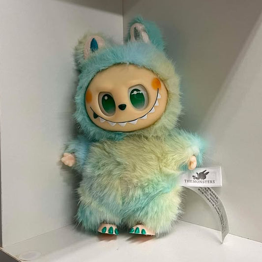 Plush toy with a fuzzy turquoise and green design on a white shelf.  It has green eyes, a green nose, a wide smile showing many teeth, a spot of blush on each cheek, and plastic hands and feet.  The feet have three toes with painted nails. 