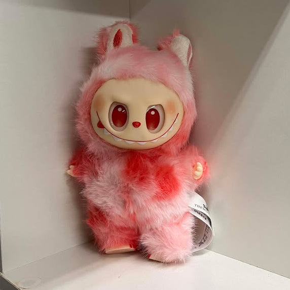 Plush toy with a fuzzy light and dark pink design on a white shelf.  It has red eyes, a red nose, a wide smile showing many teeth, a spot of blush on each cheek, and plastic hands and feet.  The feet have three toes with painted nails. 