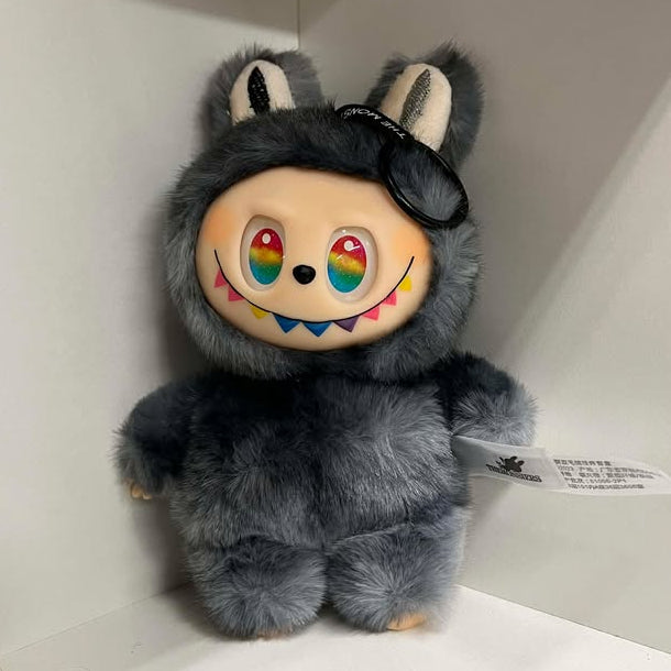 Plush toy with a fuzzy gray design on a white shelf.  It has rainbow eyes, a black nose, a wide smile showing many teeth in colors of the rainbow, a spot of blush on each cheek, and plastic hands and feet.  The feet have three toes with painted nails. 