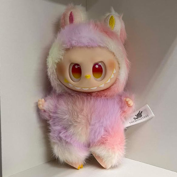 Plush toy with a fuzzy light  pink purple and white design on a white shelf.  It has red eyes, a yellow nose, a wide smile showing many teeth, a spot of blush on each cheek, and plastic hands and feet.  The feet have three toes with painted nails. 