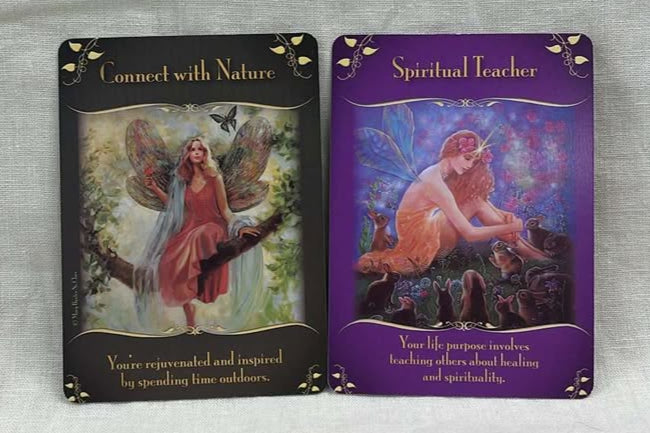 Two tarot cards on a textured surface.  One has a dark gray background, with the words "Connect with Nature" above a fair sitting on a tree limb. Below the image is the statement "You're rejuvenated and inspired by spending time outdoors."  The other has a purple background, with the words "Spiritual Teacher" above an image of a fairy sitting on the grass surrounded by small rabbits looking up at her.  Below, the card states "your life purpose involves teaching others about healing and spirituality." 