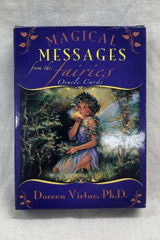 Oracle card deck cover titled 'Magical Messages from the Fairies' by Doreen Virtue on a gray background.  Deck is blue, with orange and yellow writing. In the middle is a picture of a fairy sitting in grass, with trees around her and vines made into a crown on her head and vines draped around her arms.