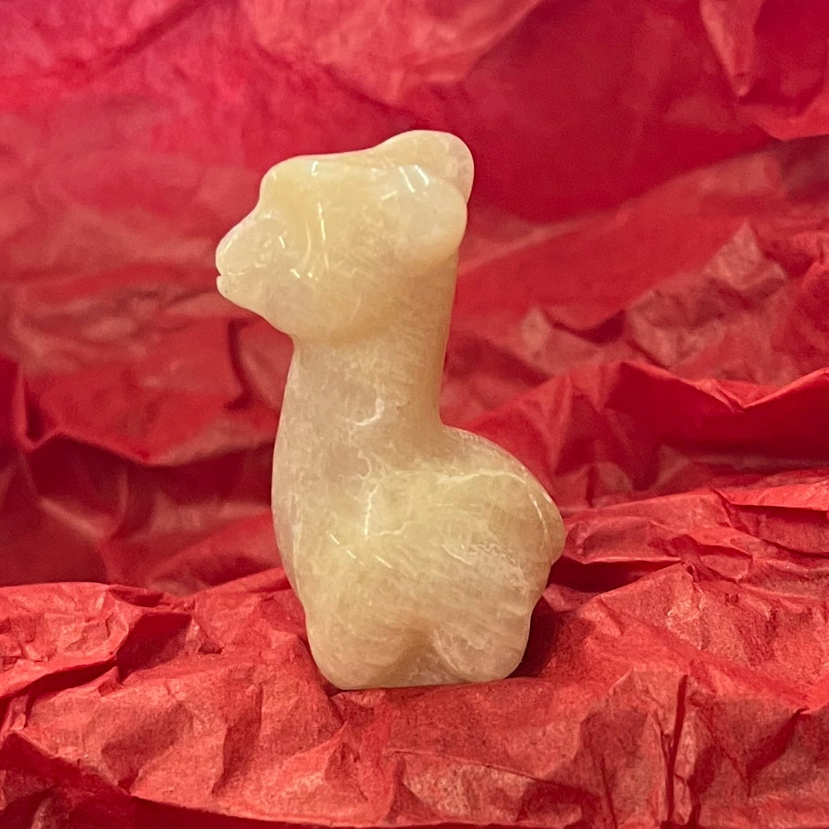 A small cream-colored carved figurine of a llama or alpaca standing upright, placed on crumpled red tissue paper. This is a side view