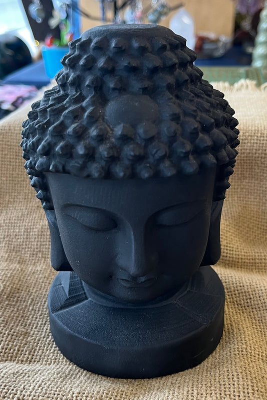 Black Buddha head figure on a burlap surface with a blurred indoor background.  The head is a dark charcoal gray, matte. 