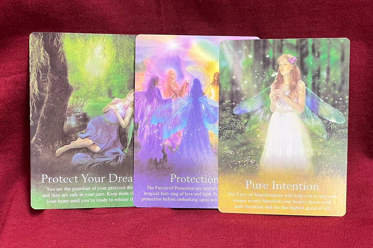 Three oracle cards with colorful fantasy illustrations of fairies are laid flat on a red fabric background. The visible card titles include 'Protect Your Dream,' 'Protection,' and 'Pure Intention.