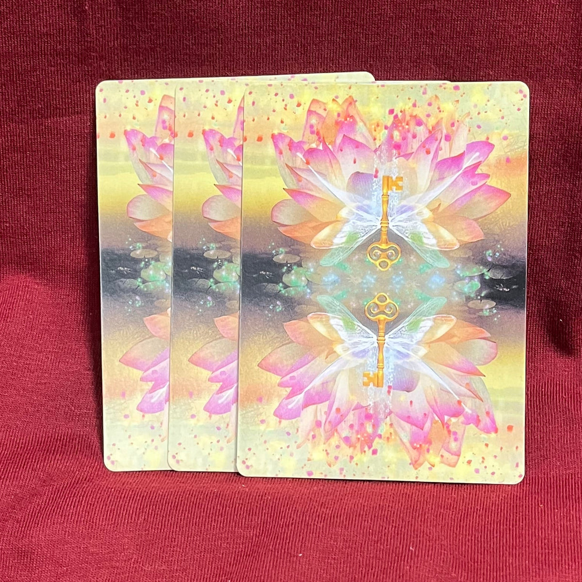 Three overlapping oracle cards with a pink and gold lotus design, resting on a dark red fabric background.