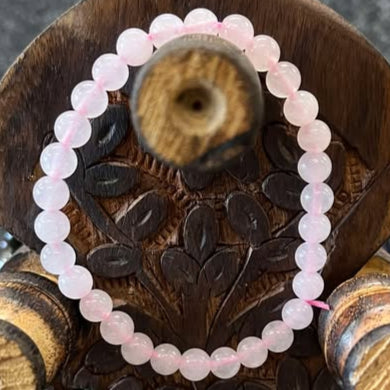 Bracelet of small, pale pink semi-translucent rose quartz beads with a light pink cord running through, on a background of brown wood. 