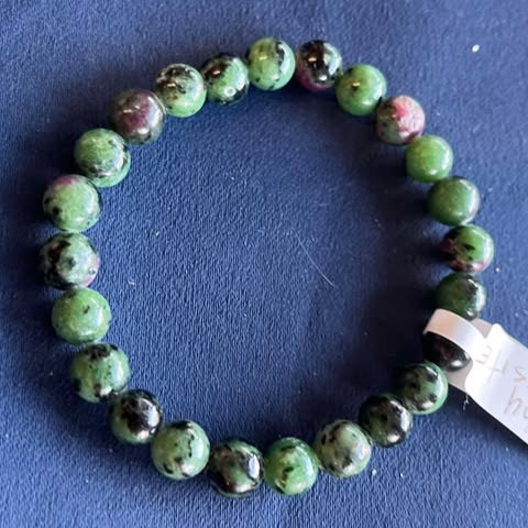 Green beaded bracelet with a tag on a blue background. Beads have black and purple interspersed as well.