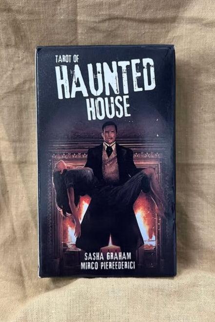 Tarot of Haunted House tarot deck front features a man in front of a fireplace, carrying a woman with long dark hair who appears to be unconscious or dead.  