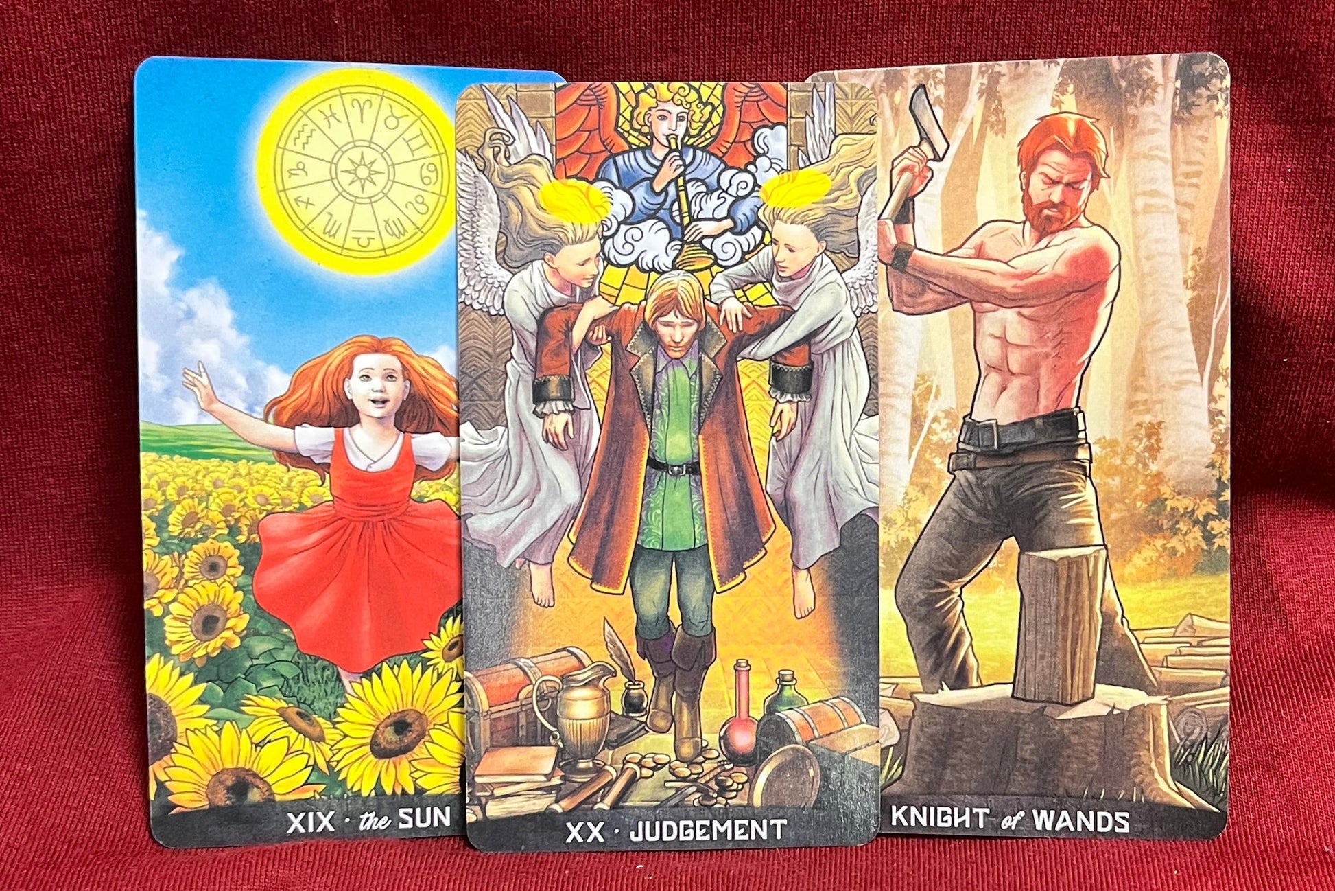 Three illustrated tarot cards laid flat on a red fabric surface. The left card shows a sunflower field with a girl in a red dress and the sun, labeled 'XIX The Sun. The middle shows a man being carried away from a man on a throne by 2 angels.  Before him is treasure and the card is labeled XX Judgement.  In the right card, a man stands with an axe in hand, ready to cut wood sitting on a stump.  The card is labeled Knight of Wands