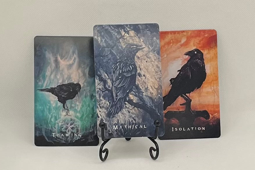 Three cards featuring watercolor-style ravens on surreal backgrounds with a word at the bottom.  One is captioned "thawing" and appears to have the raven perched on a block of ice.  Oneis captioned "mythical" and features a raven perched on a branch.  One is captioned "isolation" and features a raven  in front of a a orange textured background.  