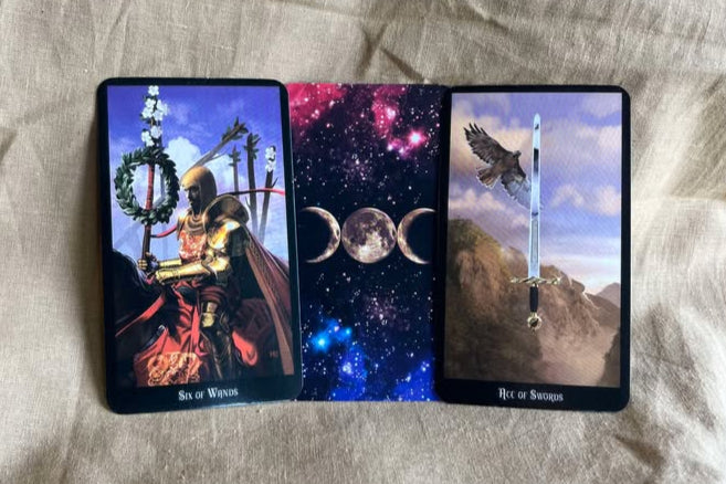 Three tarot cards displayed on a textured surface.  One features a man in armor on horseback, holding a wand with a wreath and flowers at the end.  Six of Wands. One features a card back with a solar system with stars and pink and blue gasses, with a full moon between two slivers of moon.  The third features a bird carrying a sword on a background of mountains and blue sky.  Ace of Swords