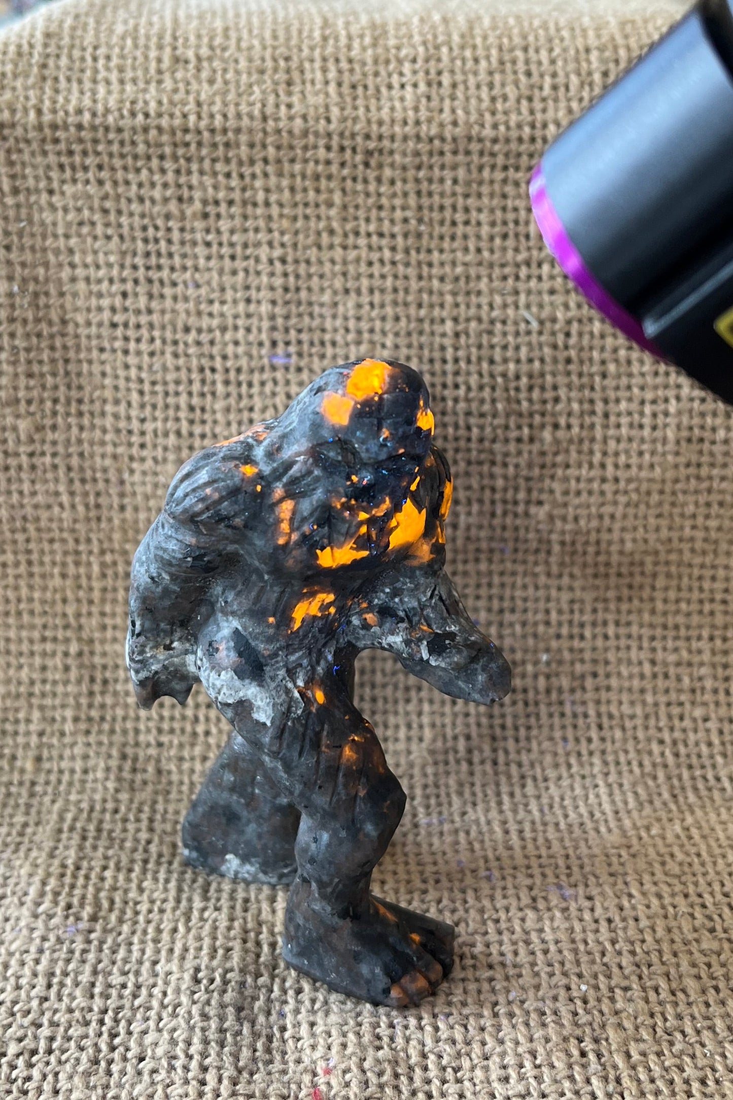 Gray black and tan crystal sasquatch on a tan textured background.  A black light is pointed at the figure and the areas in the light have spots glowing a yellowish-orange.