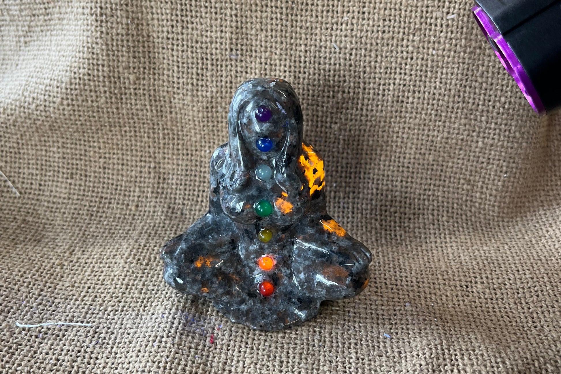 Gray, black, and tan textured stone in the shape of a woman meditating, with crystals representing the colors of the chakras running up her body.  A black light is focused on the figure and some areas have turned a yellow-ish orange where the light hit them.
