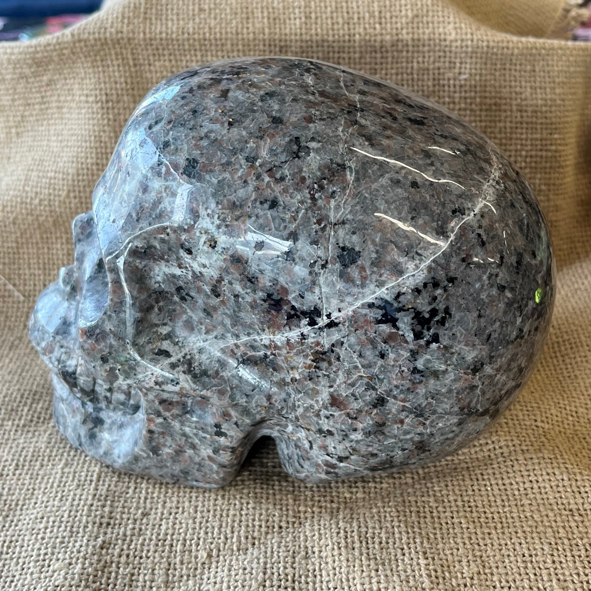 A gray and black patterned stone skull sits on on its side on a tan textured background. Some cracks are visible running through the crystal.