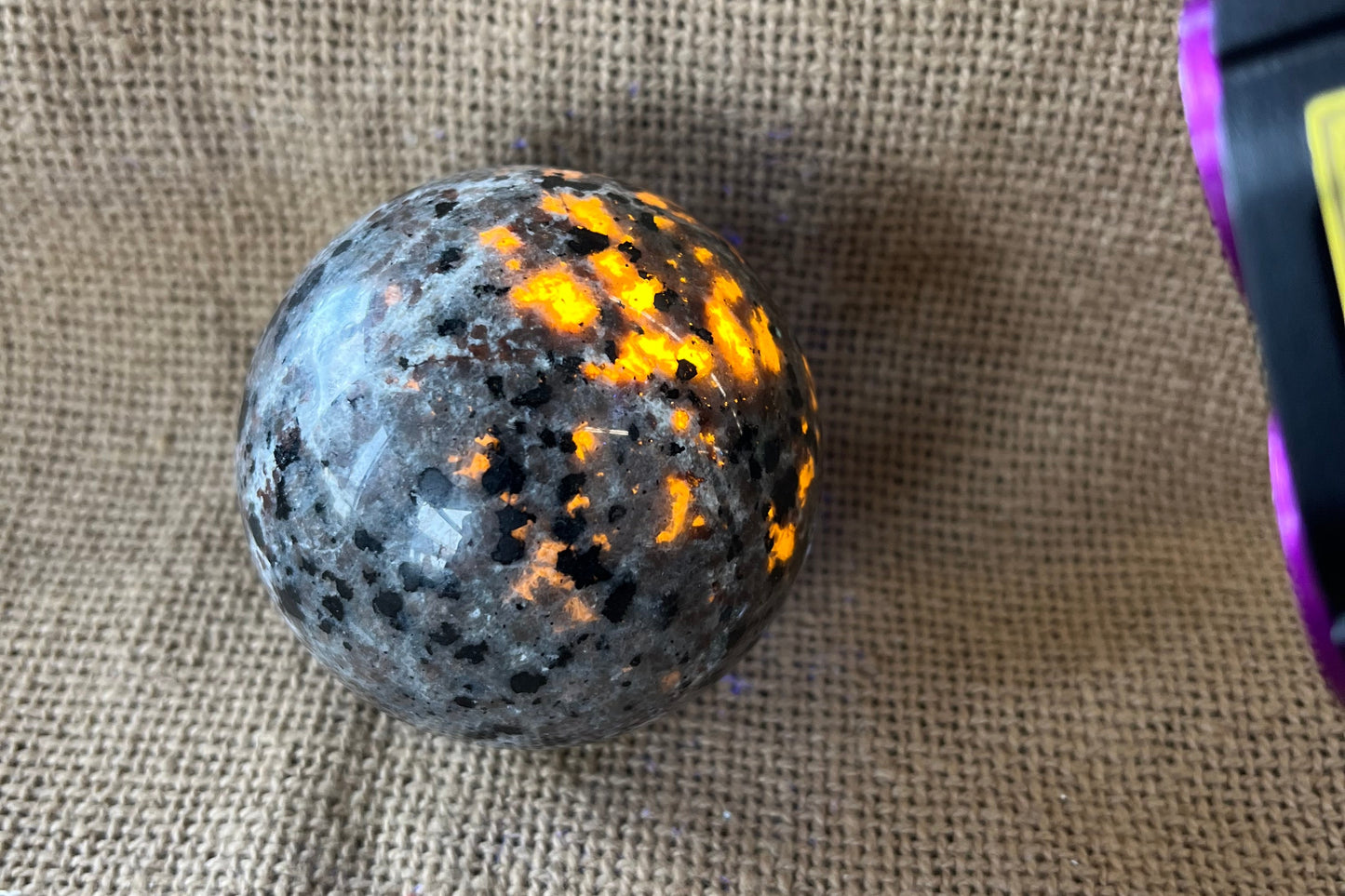 Gray black and tan crystal sphere on a tan textured background.  A black light is pointed at the sphere and the areas in the light have spots glowing a yellowish-orange.