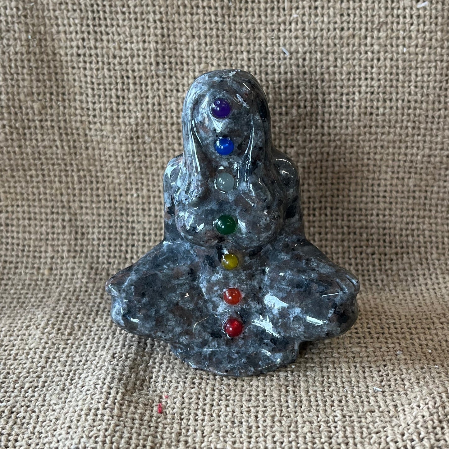 Gray, black, and tan textured stone in the shape of a woman meditating, with crystals representing the colors of the chakras running up her body. 