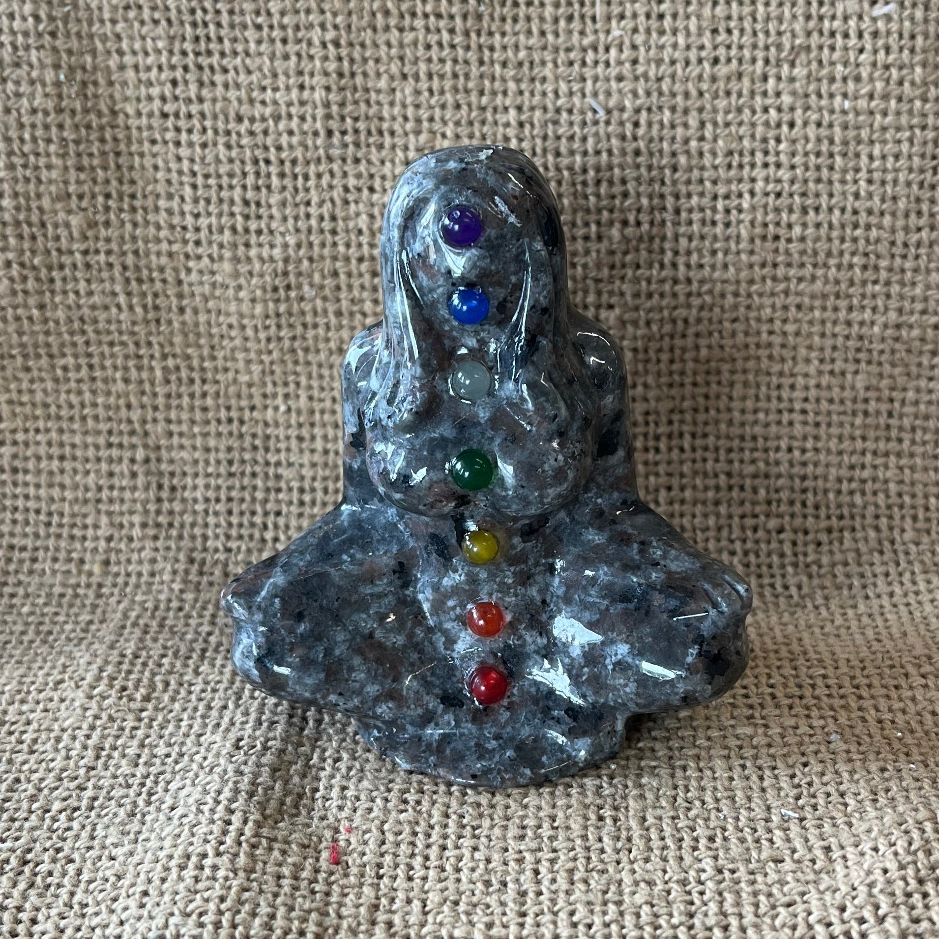 Gray, black, and tan textured stone in the shape of a woman meditating, with crystals representing the colors of the chakras running up her body. 